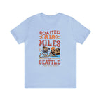 Light blue short-sleeve T-shirt displayed flat on a white background featuring a colorful graphic that reads “Roasted 818 Miles East of Seattle,” with a steaming coffee cup and mountain lake illustration on the front.