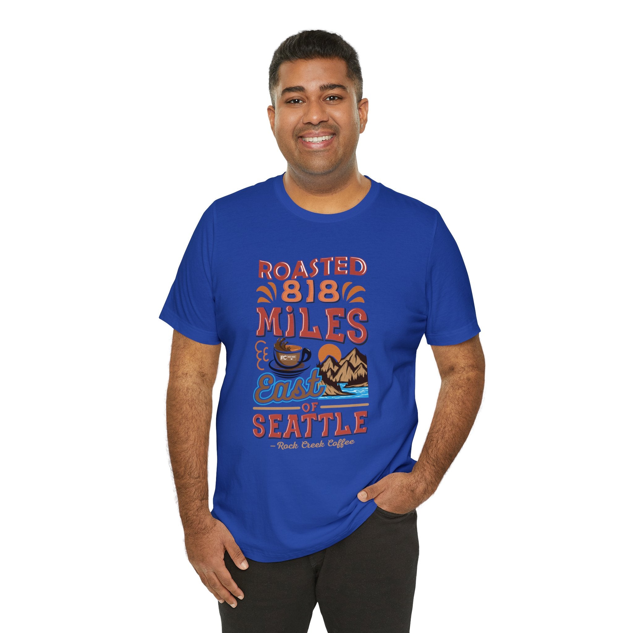 Smiling man modeling a royal blue short-sleeve T-shirt printed with a colorful graphic reading “Roasted 818 Miles East of Seattle,” featuring a steaming coffee cup and mountain lake illustration, shown against a plain white background.