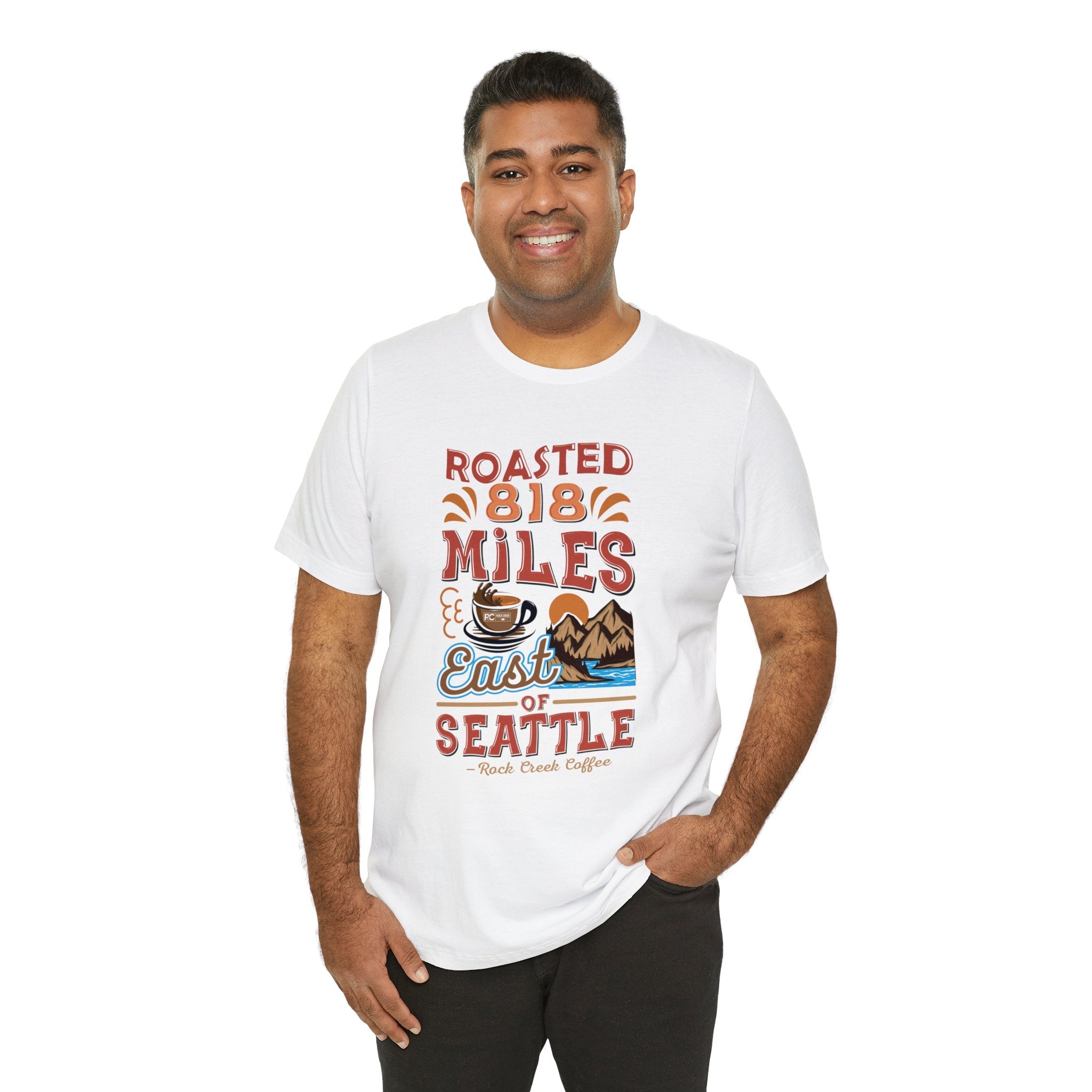 Adult man modeling a white short-sleeve T-shirt printed with a colorful coffee-themed graphic reading “Roasted 818 Miles East of Seattle” featuring a steaming coffee cup and mountain landscape, shown against a plain white background.