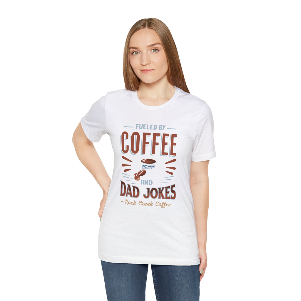 Adult woman modeling a white short-sleeve T-shirt featuring the “Fueled by Coffee and Dad Jokes” graphic with a coffee cup illustration and Rock Creek Coffee text, shown against a plain white background.
