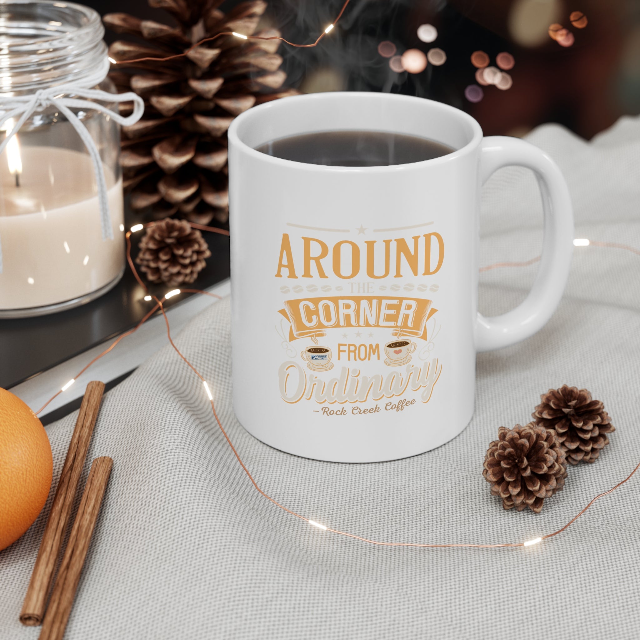 White Rock Creek Coffee mug with “Around the Corner from Ordinary” design in gold and cream lettering, filled with hot coffee and styled with pinecones, a lit candle, cinnamon sticks, an orange, and warm string lights in a cozy setting.