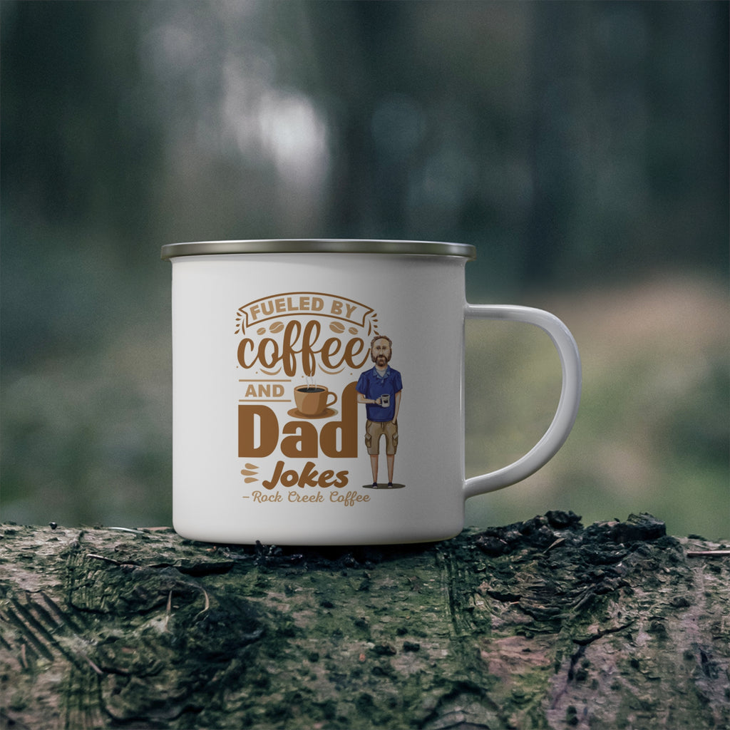 White enamel style coffee mug with silver rim and handle featuring the phrase Fueled by Coffee and Dad Jokes Rock Creek Coffee in brown lettering with a coffee cup graphic and illustration of a bearded dad holding a mug, placed on a mossy log outdoors with a blurred forest background.