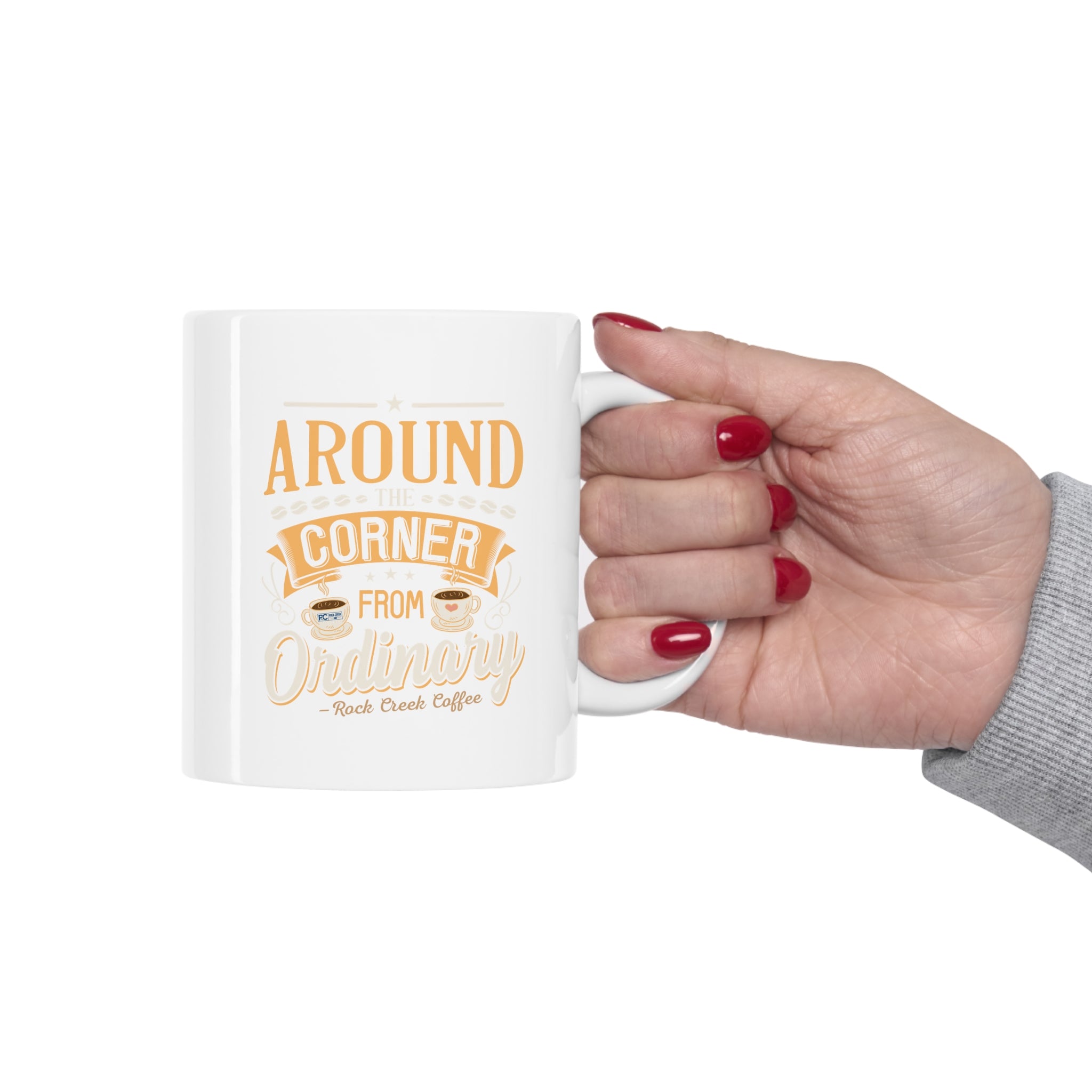 Hand with red nail polish holding a white Rock Creek Coffee mug featuring the “Around the Corner from Ordinary” design in gold and cream lettering on a clean white background.