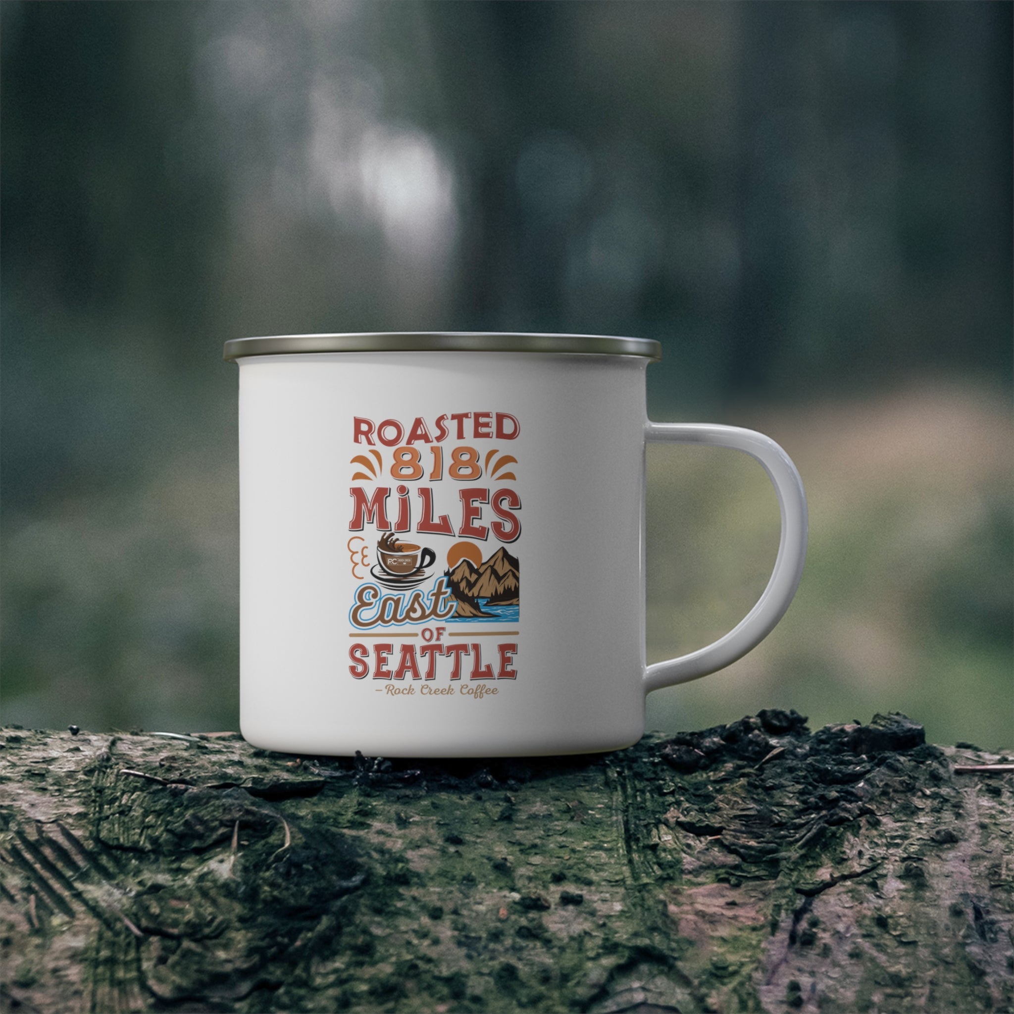 White enamel-style coffee mug with silver rim and handle featuring a colorful “Roasted 818 Miles East of Seattle – Rock Creek Coffee” design, placed on a mossy log outdoors with a blurred forest background.