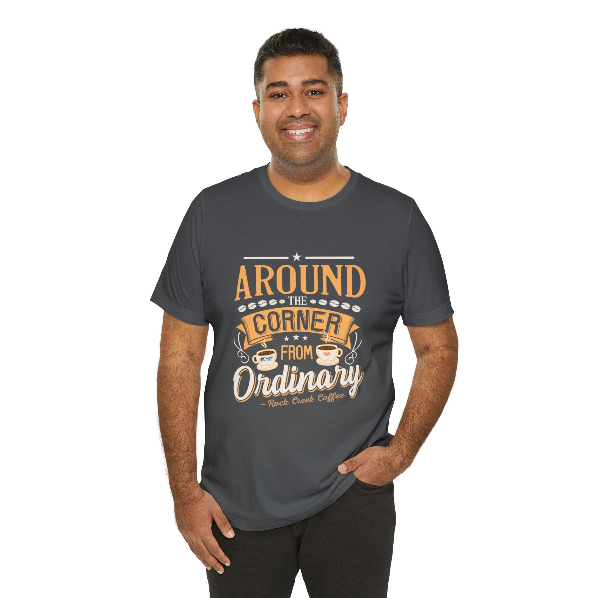 Adult man smiling while modeling an asphalt short-sleeve T-shirt featuring the “Around the Corner from Ordinary” graphic in a coffee-themed typographic design, shown against a plain white background.