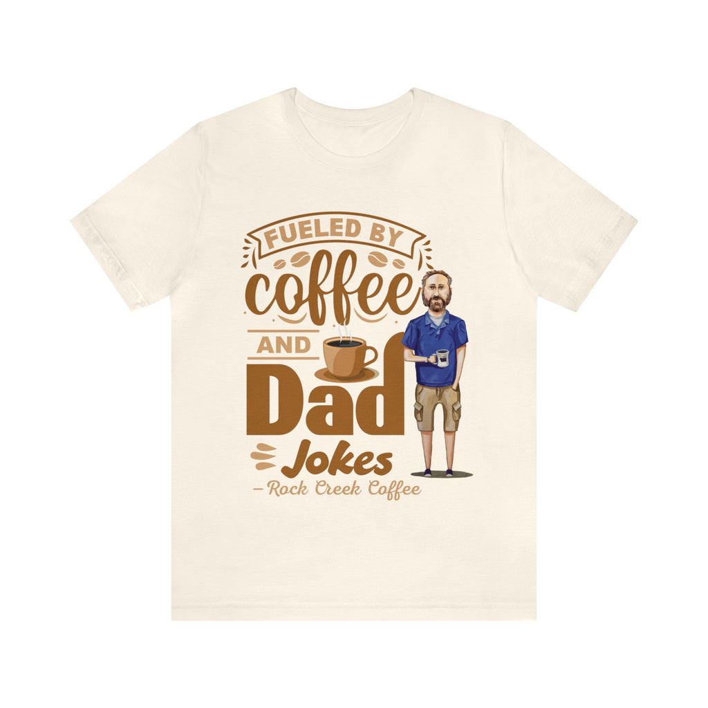 Cream short-sleeve T-shirt laid flat showing a coffee-themed graphic reading “Fueled by Coffee and Dad Jokes” with a steaming coffee cup and illustrated dad holding a mug centered on the front.