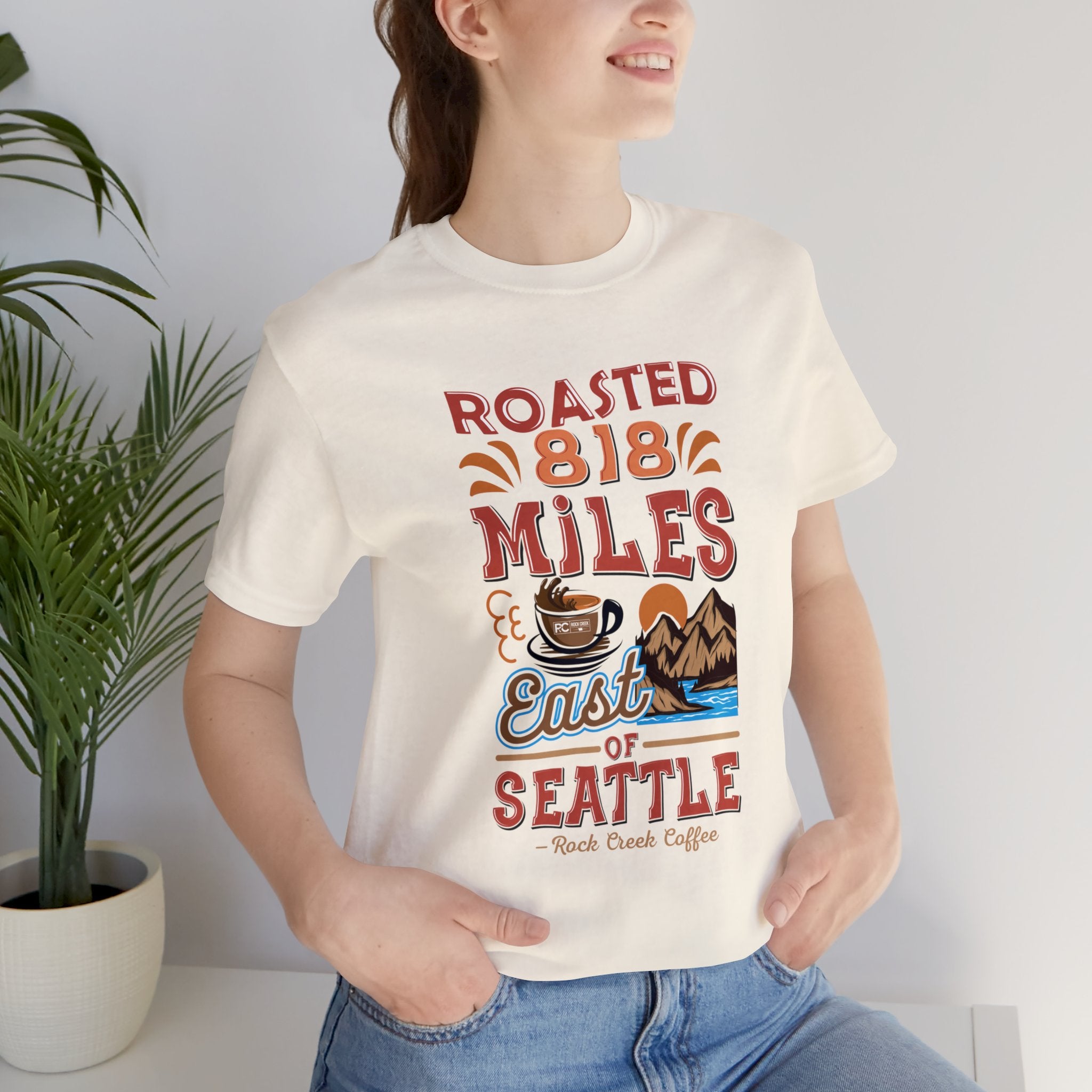 Women’s cream Seattle coffee graphic T-shirt reading “Roasted 818 Miles East of Seattle,” shown in a lifestyle photo with a potted plant and neutral indoor setting.