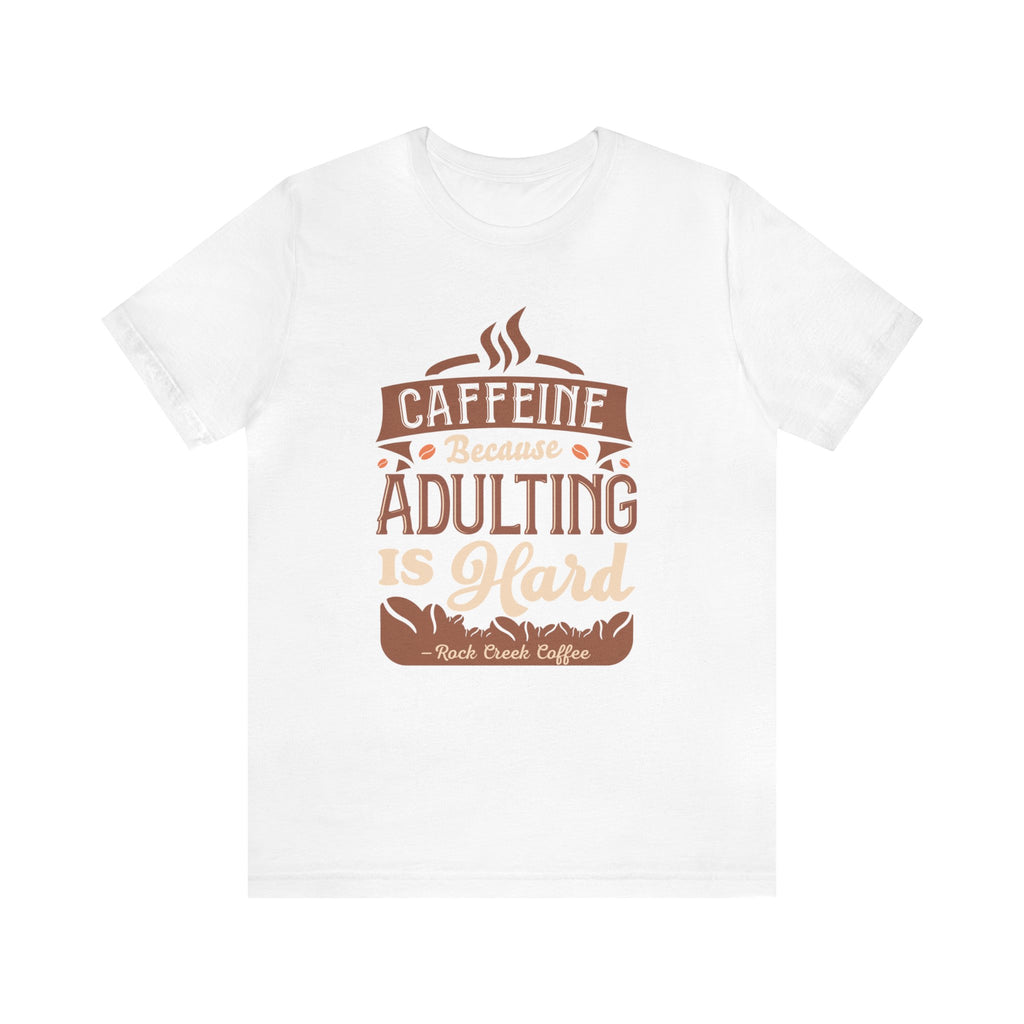 White short-sleeve T-shirt displayed flat against a plain white background, featuring the phrase “Caffeine Because Adulting Is Hard” in a stacked typographic design with coffee-inspired styling and Rock Creek Coffee text below.