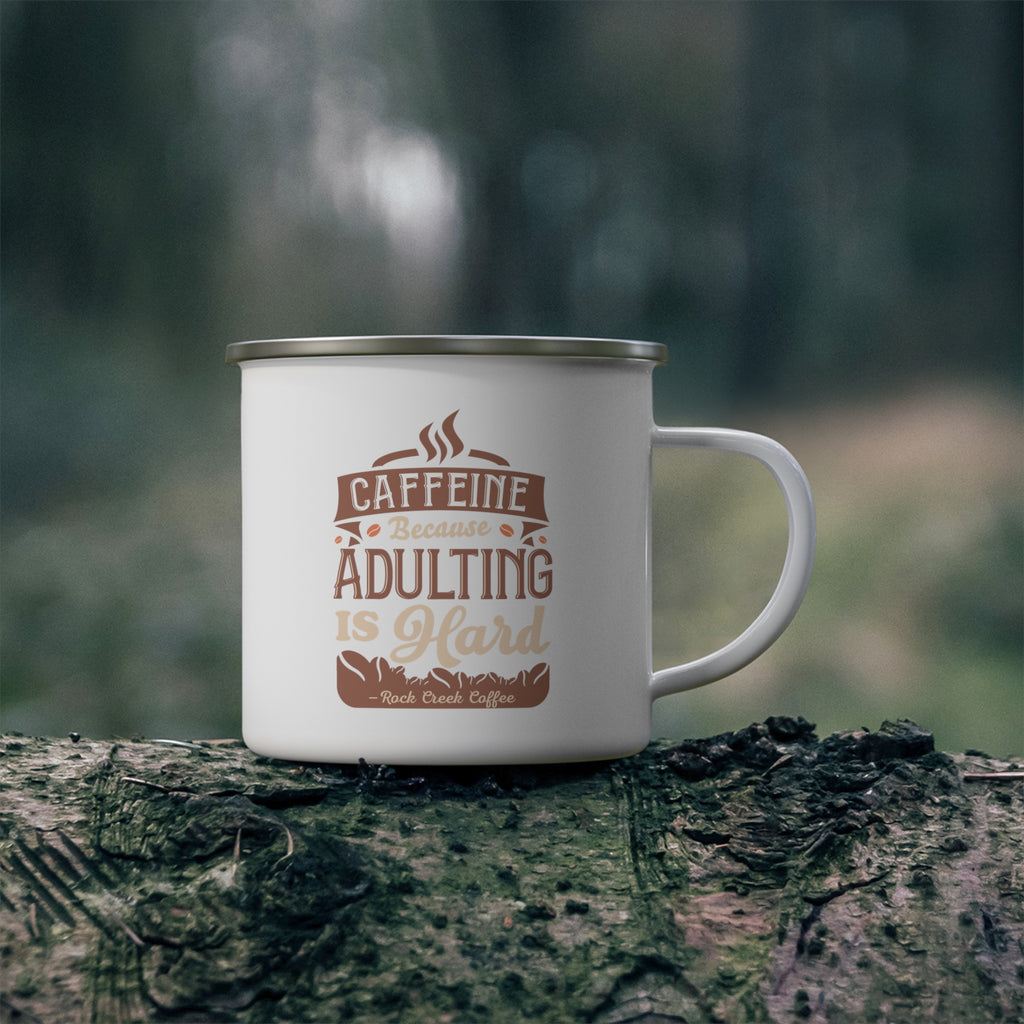 White enamel-style coffee mug with silver rim and handle, printed with “Caffeine Because Adulting Is Hard – Rock Creek Coffee,” sitting on a mossy log outdoors with a blurred forest background.