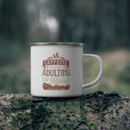 White enamel-style coffee mug with silver rim and handle, printed with “Caffeine Because Adulting Is Hard – Rock Creek Coffee,” sitting on a mossy log outdoors with a blurred forest background.