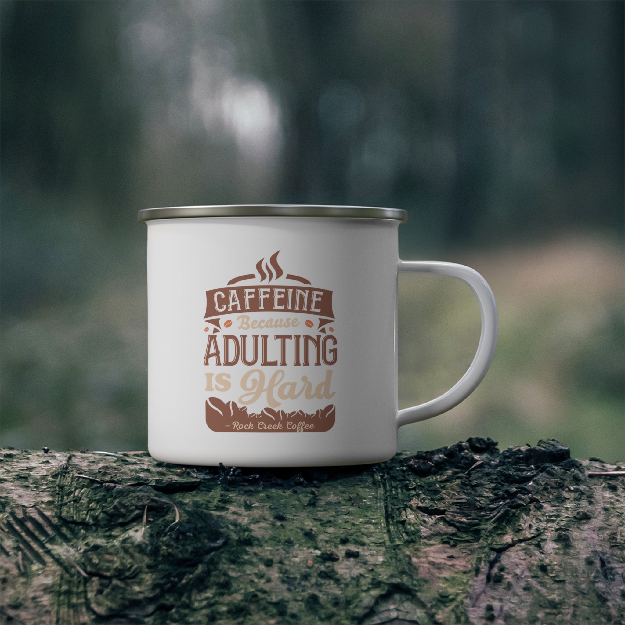 White enamel-style coffee mug with silver rim and handle, printed with “Caffeine Because Adulting Is Hard – Rock Creek Coffee,” sitting on a mossy log outdoors with a blurred forest background.
