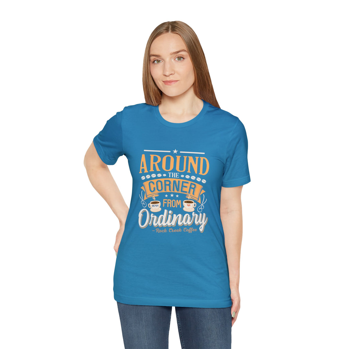 Adult woman modeling an aqua short-sleeve T-shirt featuring the “Around the Corner from Ordinary” graphic in a bold coffee-themed typographic design, shown against a plain white background.