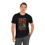 Adult man modeling a black short-sleeve T-shirt printed with a colorful graphic that reads “Roasted 818 Miles East of Seattle,” featuring a steaming coffee cup and mountain lake illustration, shown against a plain white background.