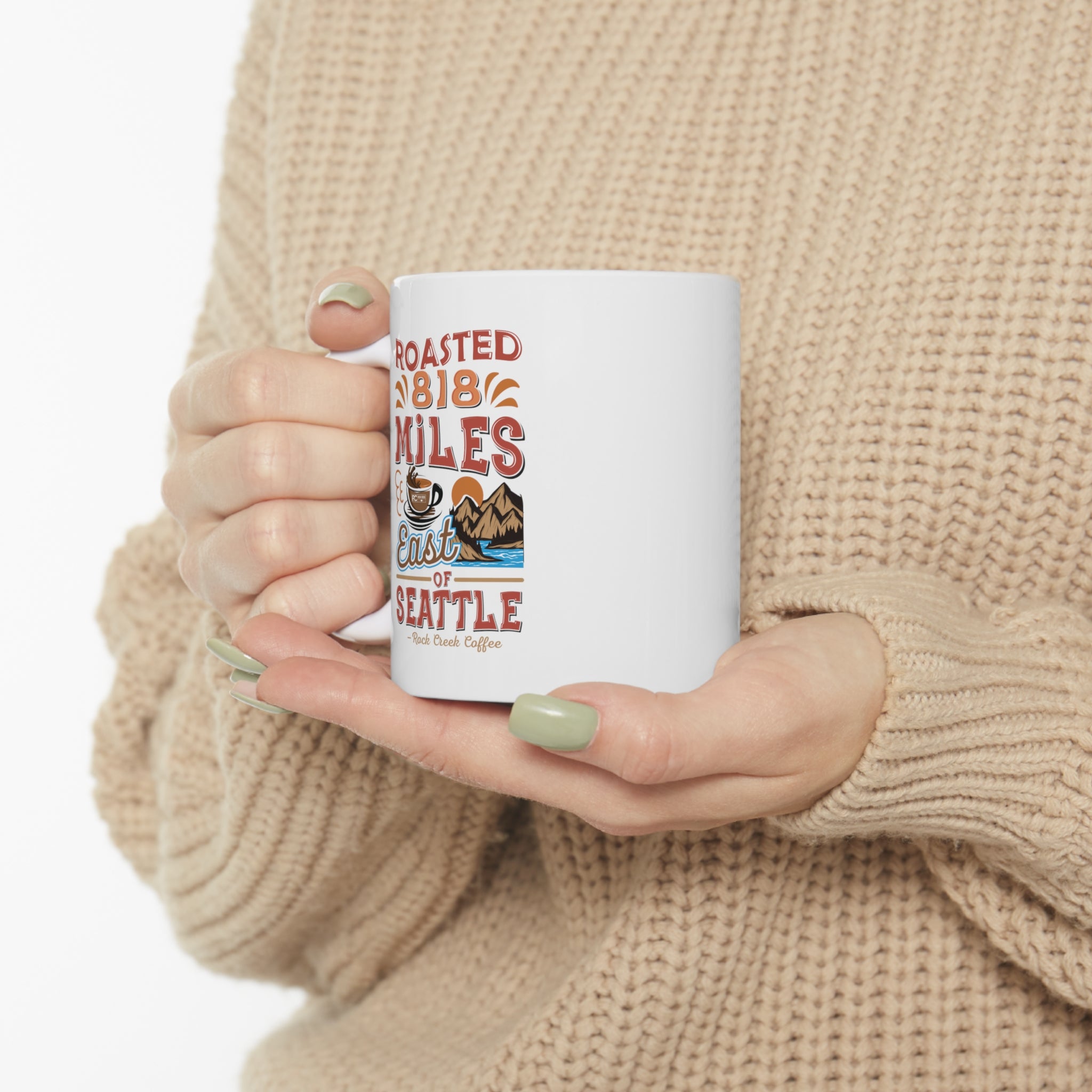 Person wearing a beige knit sweater holding a white Rock Creek Coffee mug featuring the colorful “Roasted 818 Miles East of Seattle” design.