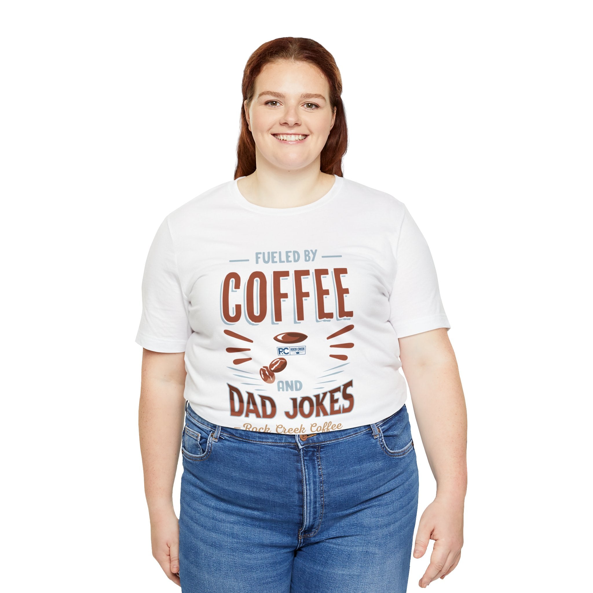 Adult woman smiling while modeling a white short-sleeve T-shirt featuring the “Fueled by Coffee and Dad Jokes” graphic with a coffee cup illustration and Rock Creek Coffee text, shown against a plain white background.