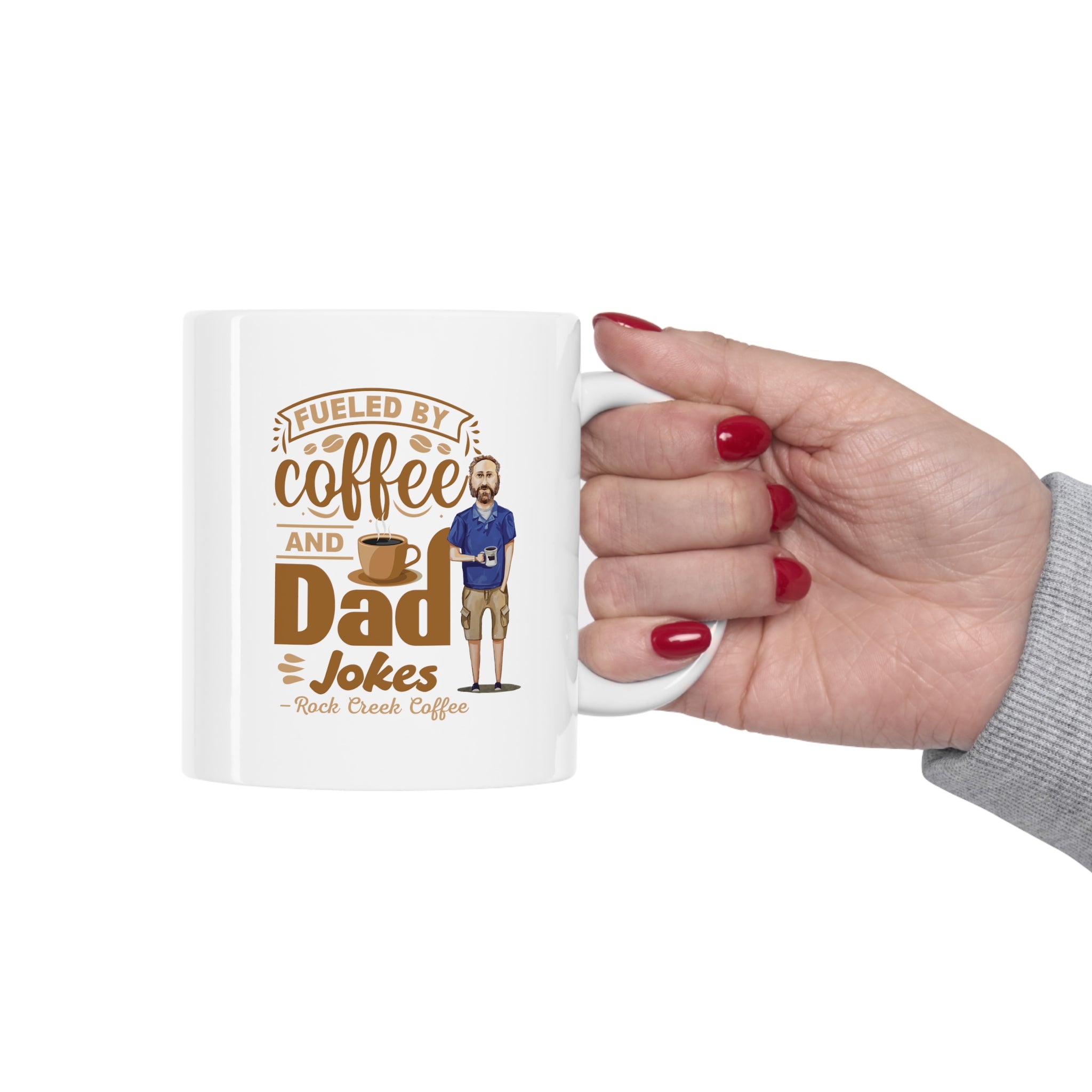 Hand holding a white ceramic mug that reads “Fueled by Coffee and Dad Jokes, Rock Creek Coffee,” featuring a cartoon illustration of a man holding a coffee cup on a clean white background.