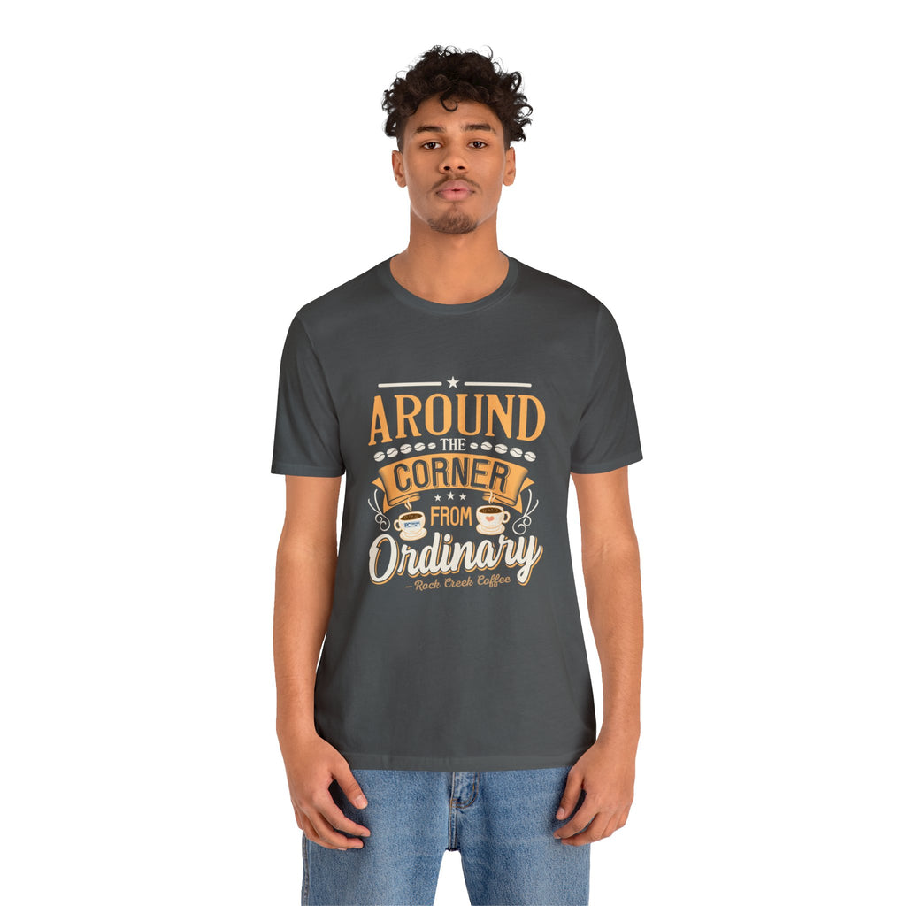 Adult man modeling an asphalt gray short-sleeve T-shirt featuring the “Around the Corner from Ordinary” graphic in a bold coffee-themed typographic design, shown against a plain white background.