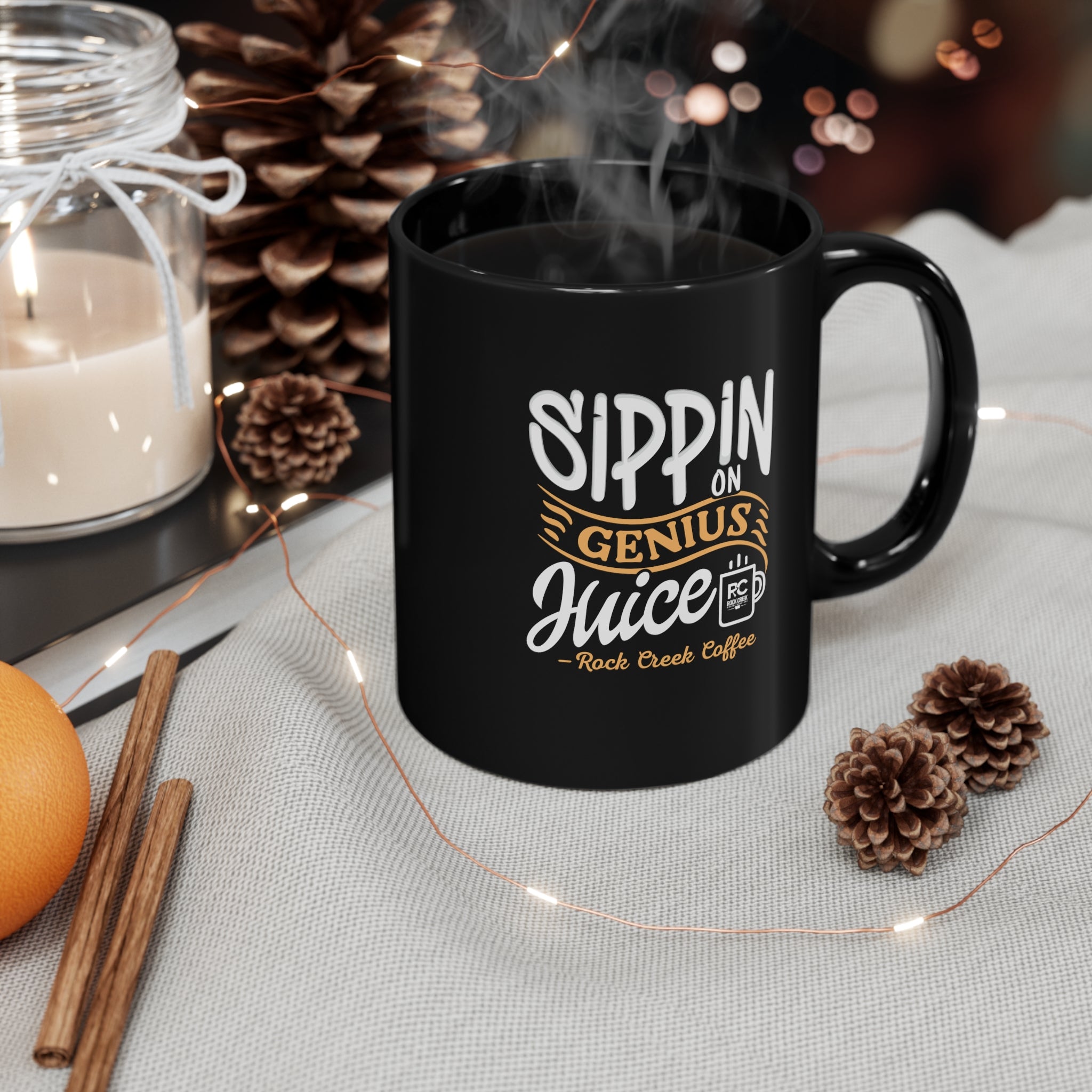 Black ceramic coffee mug with “Sippin on Genius Juice Rock Creek Coffee” design in white and gold lettering, filled with hot coffee and steam rising, displayed on a cozy tabletop with pinecones, a lit candle, cinnamon sticks, and soft string lights in the background.
