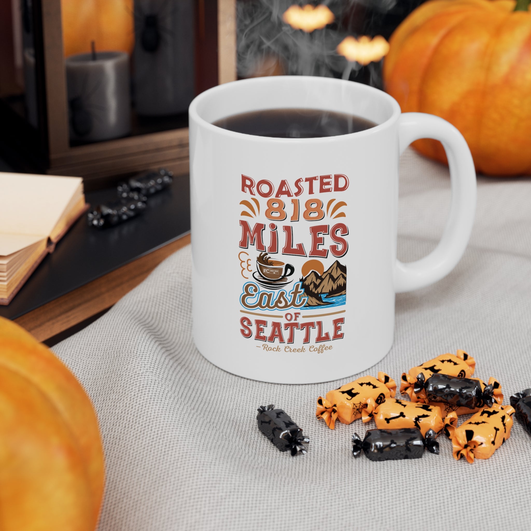 White Rock Creek Coffee mug featuring the colorful “Roasted 818 Miles East of Seattle” design, filled with hot coffee and styled with pumpkins and Halloween candies on a cozy fall themed tabletop.