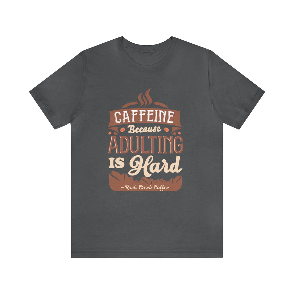 Asphalt short-sleeve T-shirt displayed flat against a plain white background, featuring the “Caffeine Because Adulting Is Hard” graphic in a stacked typographic design with Rock Creek Coffee branding below.