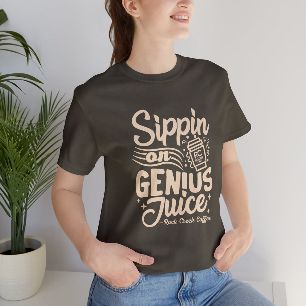 Smiling woman wearing an olive green short-sleeve T-shirt printed with the slogan “Sippin on Genius Juice” and a stylized coffee to-go cup graphic, seated casually indoors with a potted plant in the background.