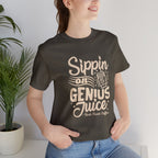 Smiling woman wearing an olive green short-sleeve T-shirt printed with the slogan “Sippin on Genius Juice” and a stylized coffee to-go cup graphic, seated casually indoors with a potted plant in the background.