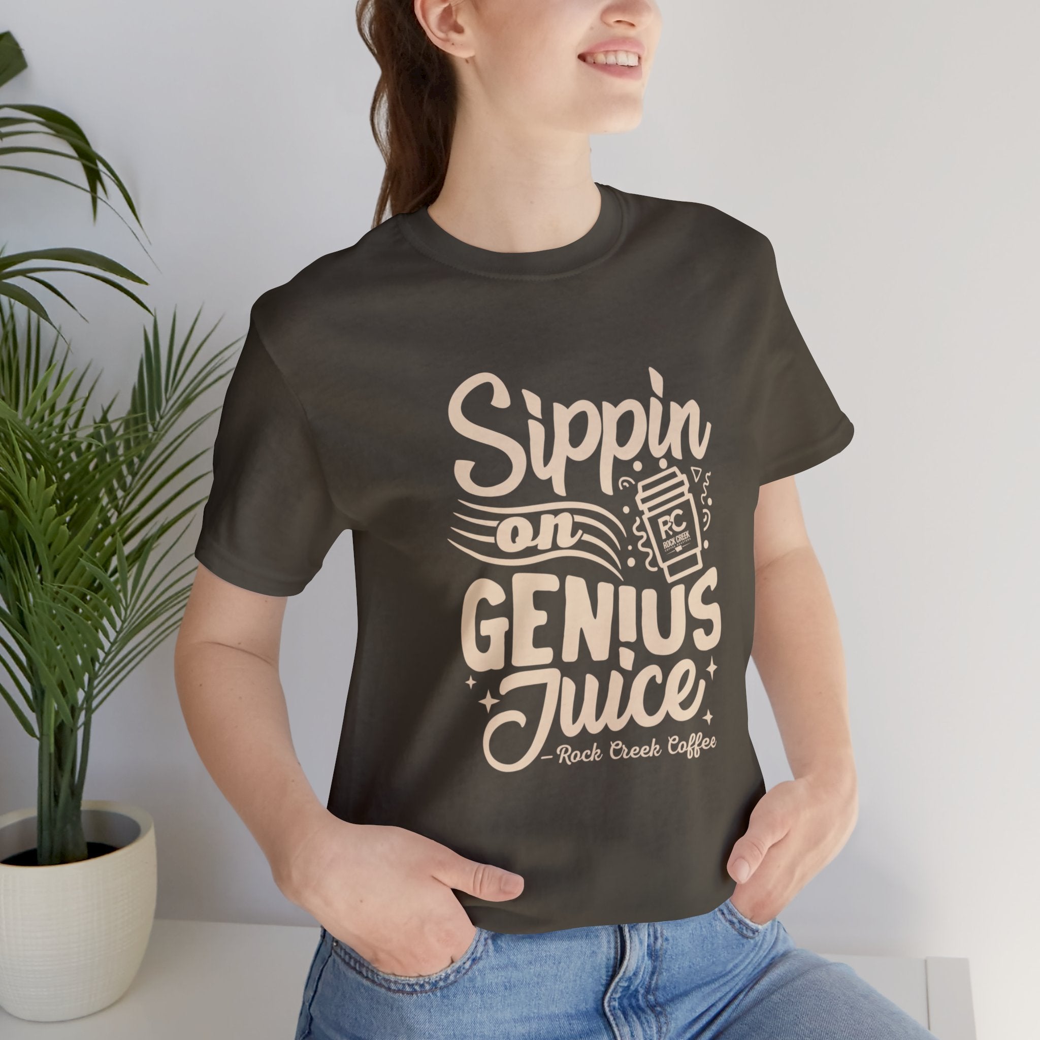 Smiling woman wearing an olive green short-sleeve T-shirt printed with the slogan “Sippin on Genius Juice” and a stylized coffee to-go cup graphic, seated casually indoors with a potted plant in the background.