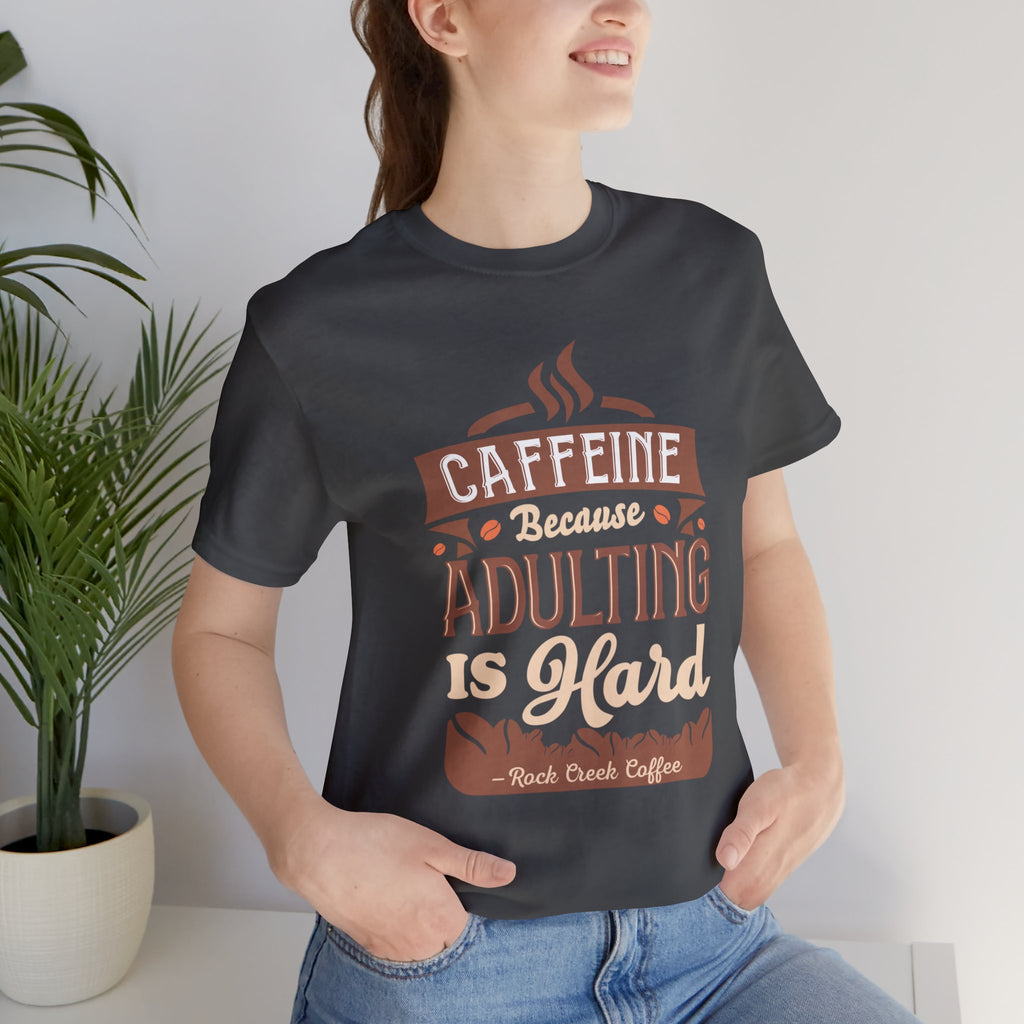 Adult woman modeling an asphalt short-sleeve T-shirt featuring the “Caffeine Because Adulting Is Hard” graphic in a stacked typographic design, photographed indoors beside a potted plant against a light neutral background.