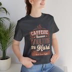 Adult woman modeling an asphalt short-sleeve T-shirt featuring the “Caffeine Because Adulting Is Hard” graphic in a stacked typographic design, photographed indoors beside a potted plant against a light neutral background.