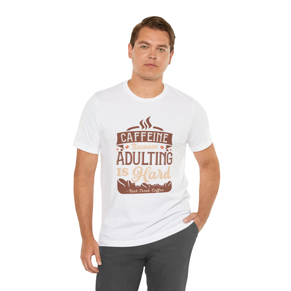 Adult man posing with one hand in his pocket while modeling a white short-sleeve T-shirt featuring the “Caffeine Because Adulting Is Hard” graphic in a stacked typographic design, shown against a plain white background.