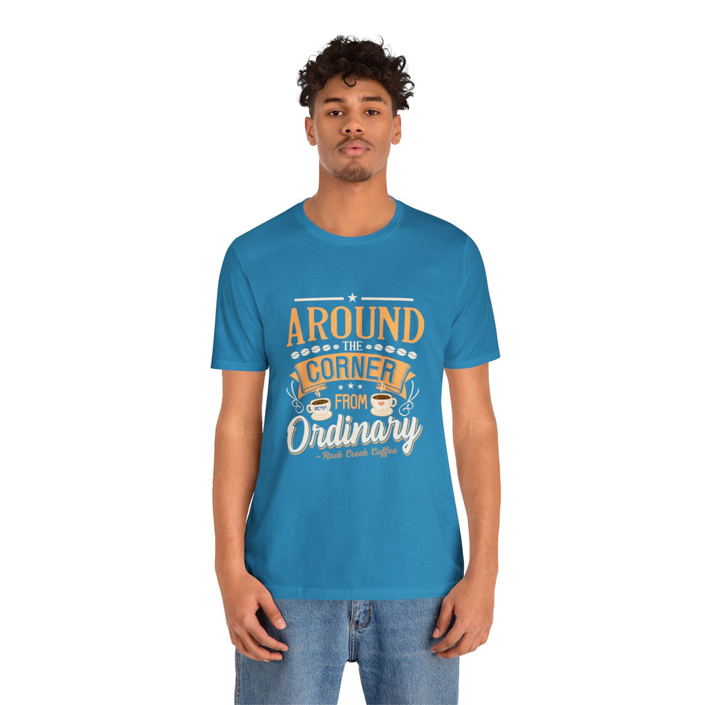 Adult man modeling an aqua short-sleeve T-shirt featuring the phrase “Around the Corner from Ordinary” in a bold coffee-themed typographic design, shown against a plain white background.