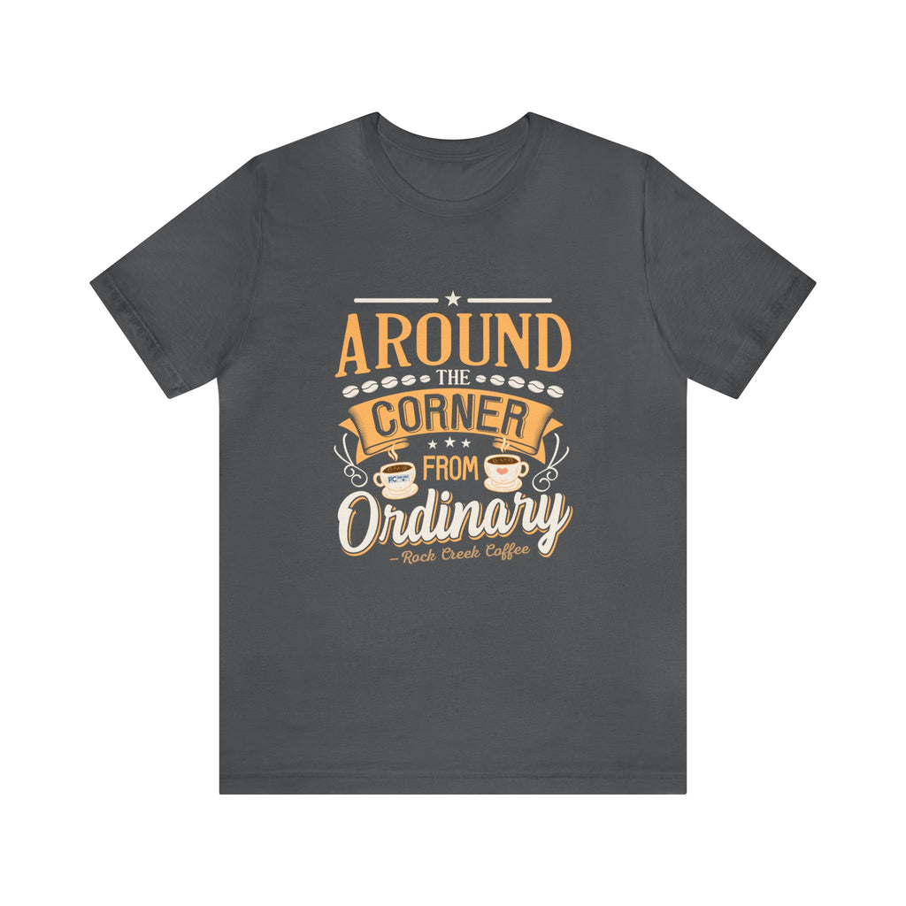 Asphalt gray short-sleeve T-shirt displayed flat against a plain white background, featuring the phrase “Around the Corner from Ordinary” in a bold coffee-themed typographic design with Rock Creek Coffee branding below.