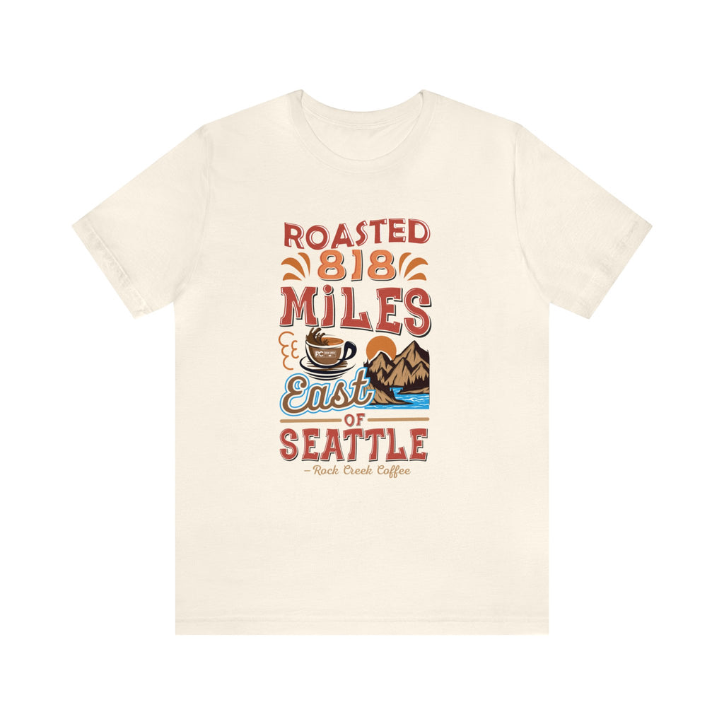 Cream short-sleeve T-shirt featuring a colorful “Roasted 818 Miles East of Seattle” graphic with a steaming coffee cup and mountain lake illustration, displayed against a plain white background.