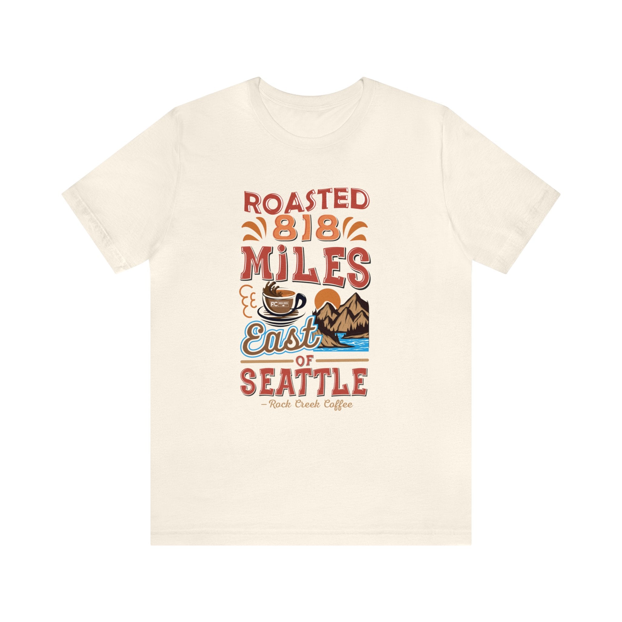 Cream short-sleeve T-shirt featuring a colorful “Roasted 818 Miles East of Seattle” graphic with a steaming coffee cup and mountain lake illustration, displayed against a plain white background.