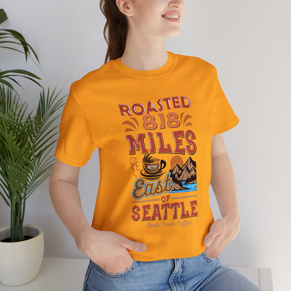 Adult woman modeling a golden yellow short-sleeve T-shirt printed with a colorful graphic that reads “Roasted 818 Miles East of Seattle,” featuring a steaming coffee cup and mountain lake illustration, shown in an indoor setting with a potted plant and light background.