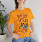 Adult woman modeling a golden yellow short-sleeve T-shirt printed with a colorful graphic that reads “Roasted 818 Miles East of Seattle,” featuring a steaming coffee cup and mountain lake illustration, shown in an indoor setting with a potted plant and light background.