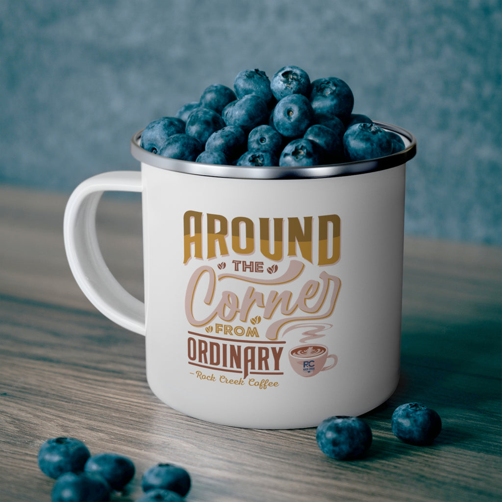 White enamel style coffee mug with silver rim filled with fresh blueberries, featuring the phrase Around the Corner from Ordinary Rock Creek Coffee in tan and brown lettering with a small latte cup illustration, set on a wooden surface with a blue textured background.