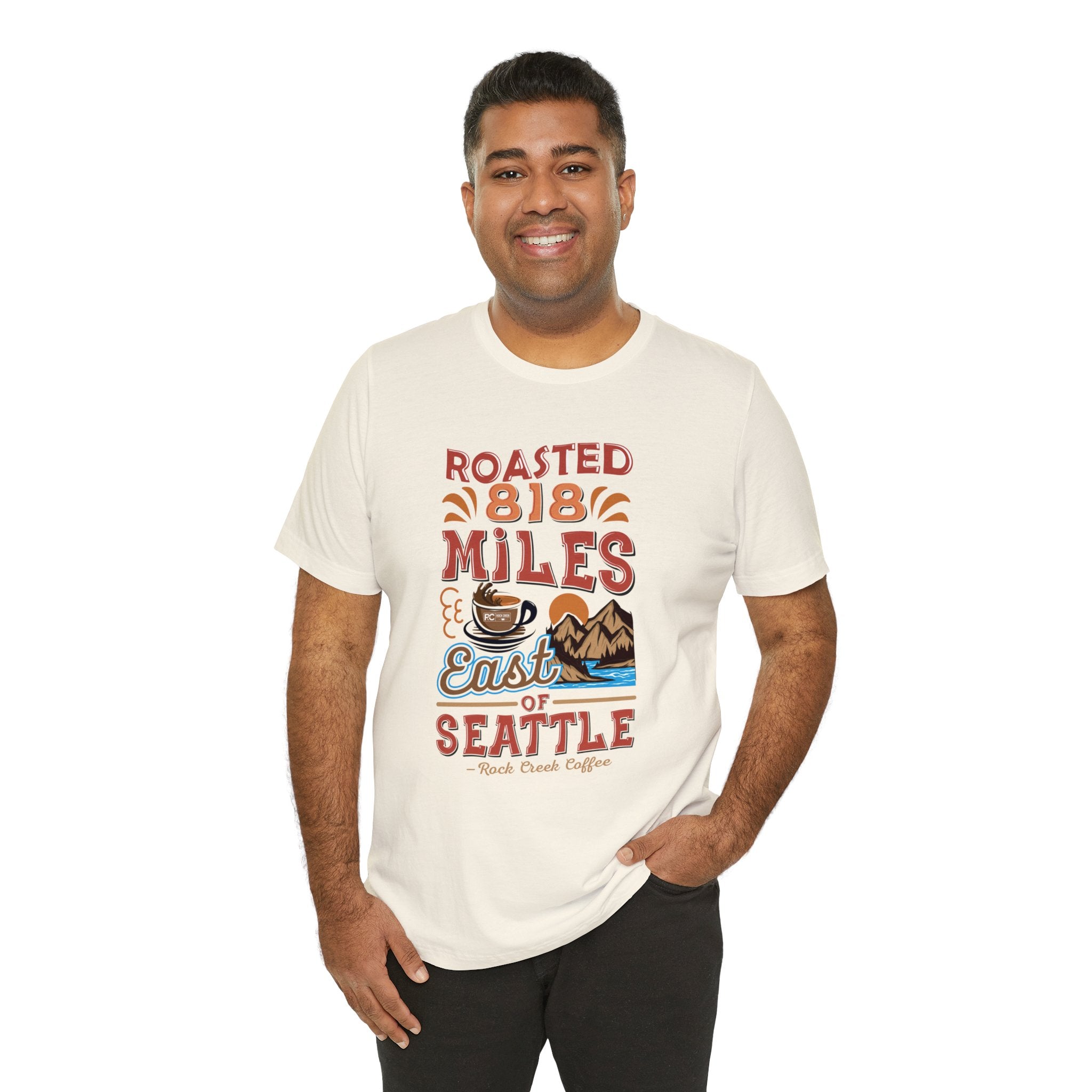 Smiling man modeling a cream short-sleeve T-shirt printed with a colorful graphic reading “Roasted 818 Miles East of Seattle,” featuring a steaming coffee cup and scenic mountain lake illustration, shown against a plain white background.