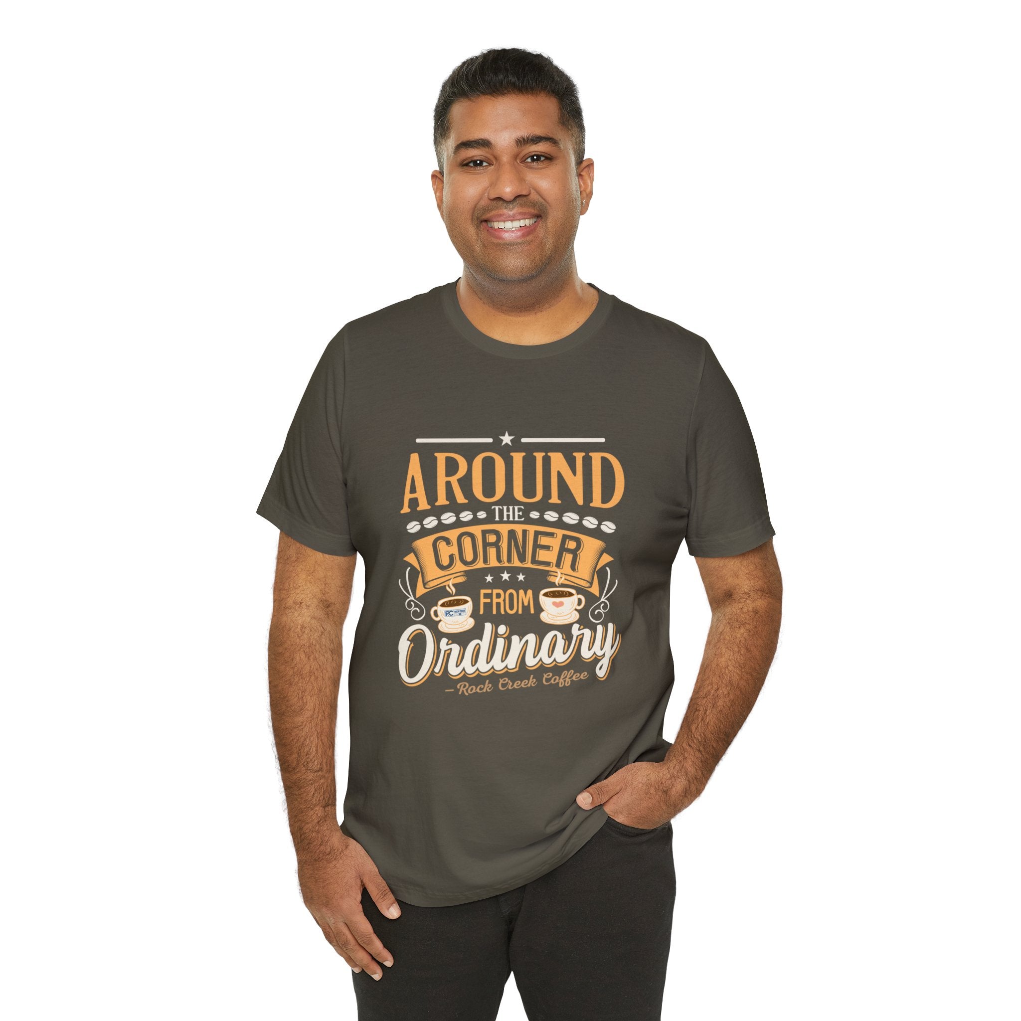 Adult man smiling while modeling army colored short-sleeve T-shirt featuring the phrase “Around the Corner from Ordinary” in a bold typographic coffee-themed design, shown against a plain white background.