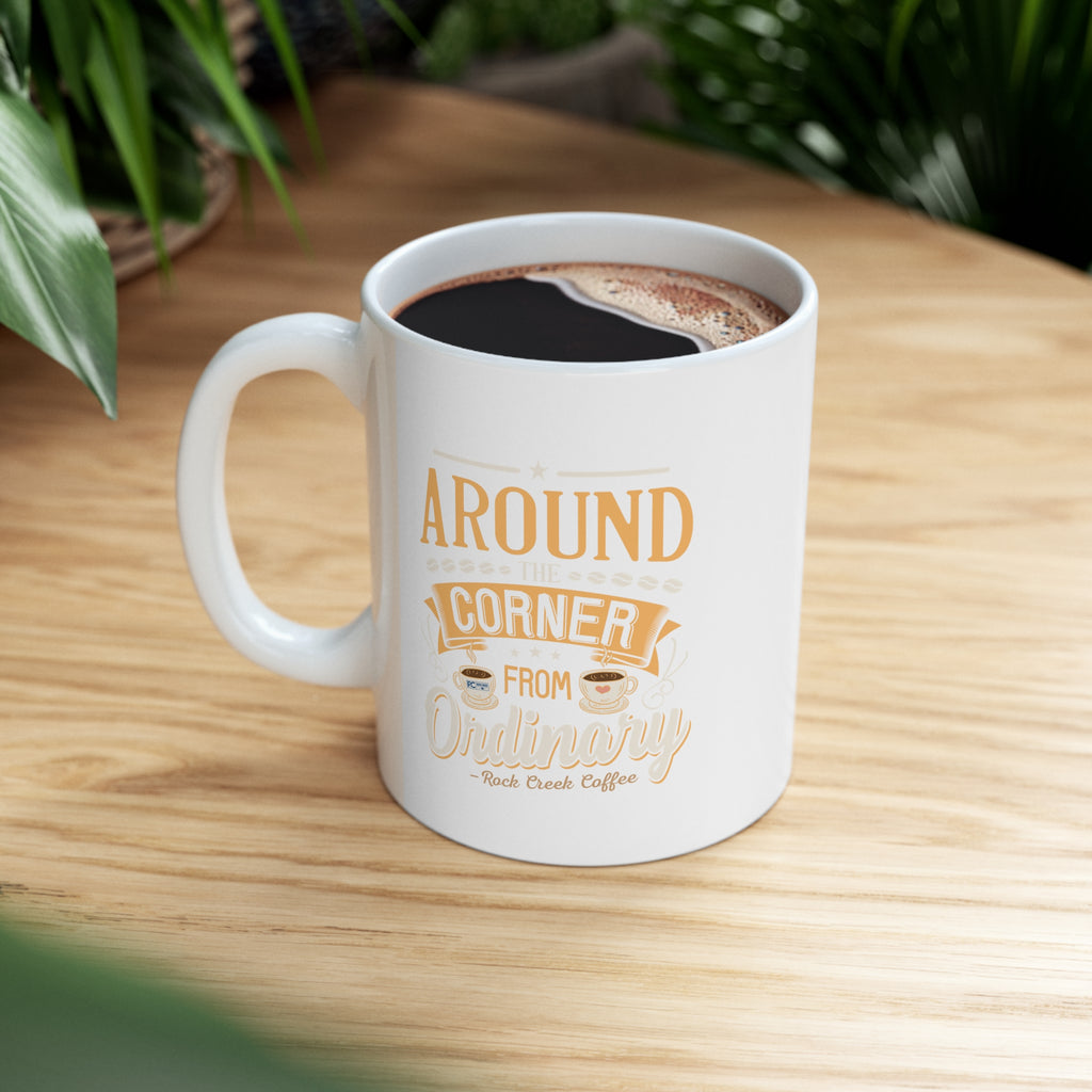 White Rock Creek Coffee mug with “Around the Corner from Ordinary” design in gold and cream lettering, filled with freshly brewed coffee and placed on a wooden table with green plants in the background.