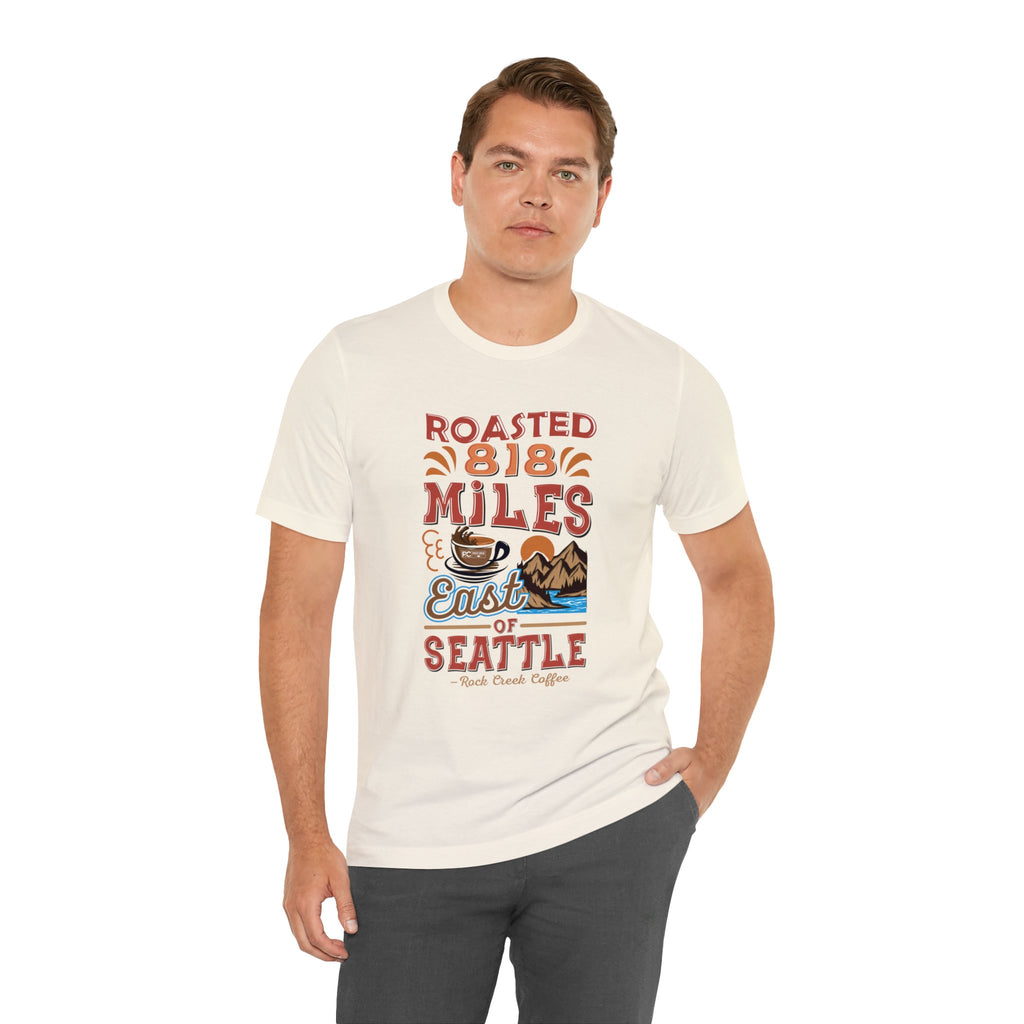 Adult man modeling a cream short-sleeve T-shirt printed with a colorful graphic that reads “Roasted 818 Miles East of Seattle,” featuring a steaming coffee cup and mountain lake illustration, shown against a plain white background.