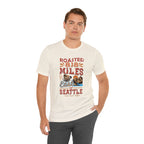 Adult man modeling a cream short-sleeve T-shirt printed with a colorful graphic that reads “Roasted 818 Miles East of Seattle,” featuring a steaming coffee cup and mountain lake illustration, shown against a plain white background.