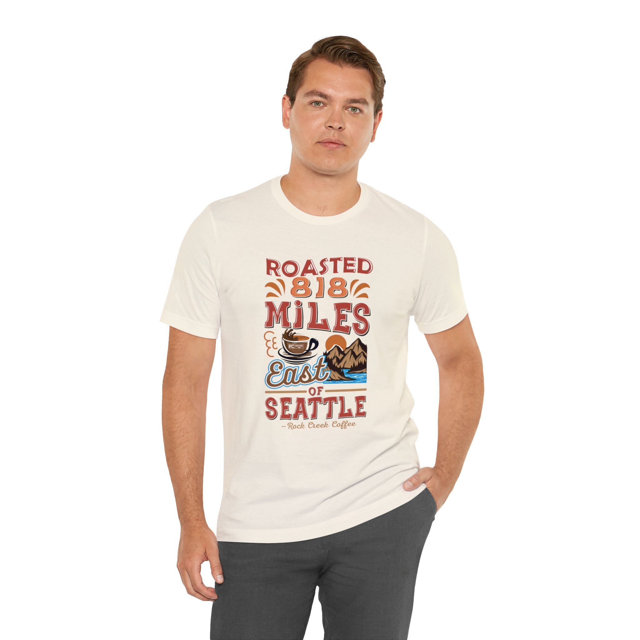 Adult man modeling a cream short-sleeve T-shirt printed with a colorful graphic that reads “Roasted 818 Miles East of Seattle,” featuring a steaming coffee cup and mountain lake illustration, shown against a plain white background.