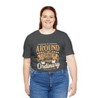 Adult woman smiling while modeling an asphalt gray short-sleeve T-shirt featuring the “Around the Corner from Ordinary” graphic in a bold coffee-themed typographic design, shown against a plain white background.