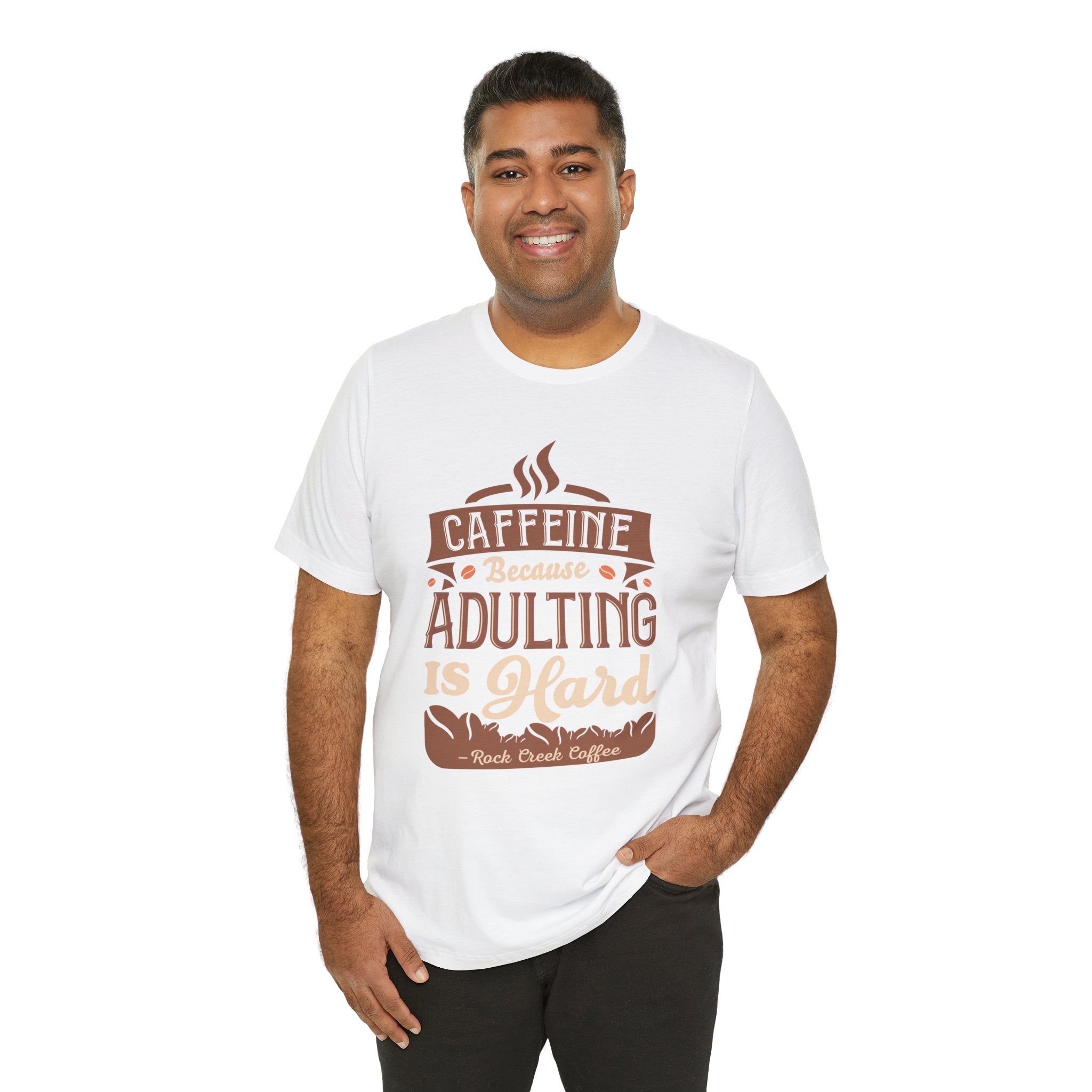 Adult man smiling while modeling a white short-sleeve T-shirt printed with the phrase “Caffeine Because Adulting Is Hard” in a stacked typographic design, shown against a plain white background.