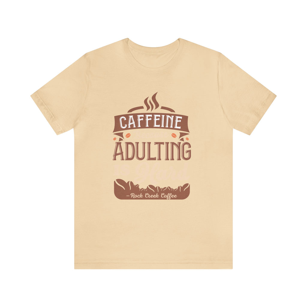 Soft cream short-sleeve T-shirt displayed flat against a plain white background, featuring the “Caffeine Because Adulting Is Hard” graphic in a stacked typographic design with Rock Creek Coffee branding below.