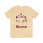 Soft cream short-sleeve T-shirt displayed flat against a plain white background, featuring the “Caffeine Because Adulting Is Hard” graphic in a stacked typographic design with Rock Creek Coffee branding below.