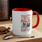 White ceramic coffee mug with a red interior and handle featuring the Roasted 818 Miles East of Seattle Rock Creek Coffee design, displayed on a wooden surface beside a spoon, coffee beans, and a chocolate macaron with a coffee grinder in the background.