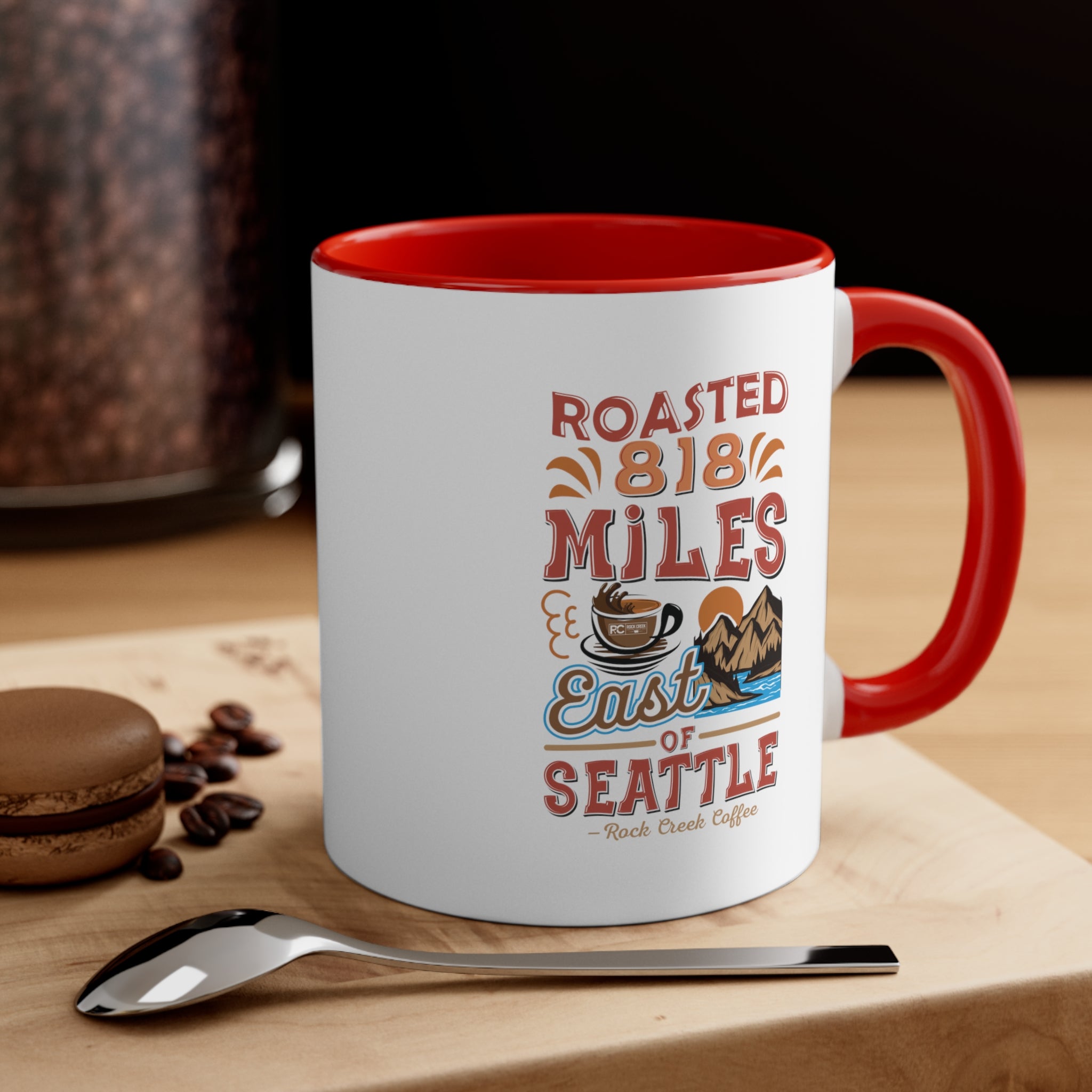 White ceramic coffee mug with a red interior and handle featuring the Roasted 818 Miles East of Seattle Rock Creek Coffee design, displayed on a wooden surface beside a spoon, coffee beans, and a chocolate macaron with a coffee grinder in the background.
