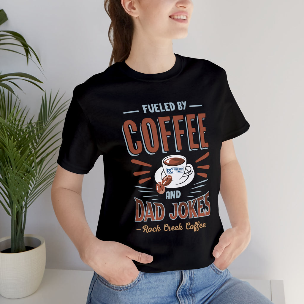 Adult woman modeling a black short-sleeve T-shirt featuring the “Fueled by Coffee and Dad Jokes” graphic with a coffee cup illustration and Rock Creek Coffee text, photographed indoors beside a potted plant against a light background.